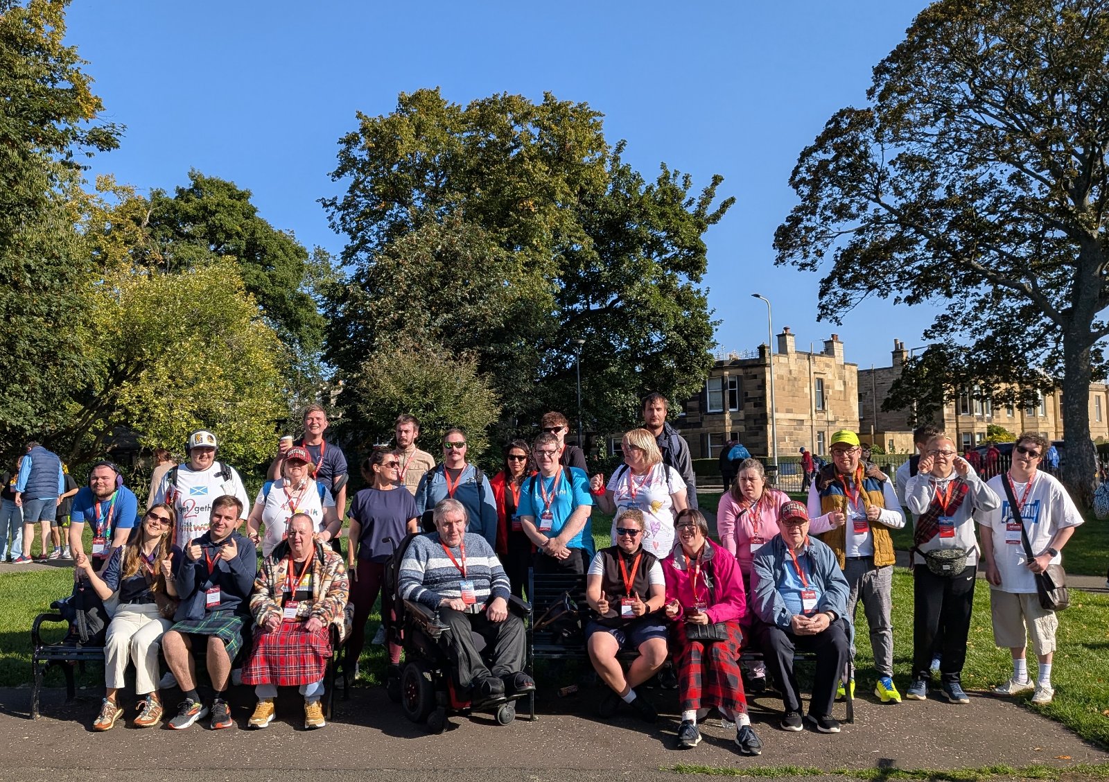 get2gether members at the start of the Kiltwalk, all standing in the park in  line and looking at the camera. It's a sunny day with trees and buildings in the backgrouns.